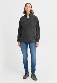 Pullover in pile grigio scuro con zip a metà, tasche frontali e un accento del logo. Abbinato a jeans blu e sneaker grigie. Texture liscia.