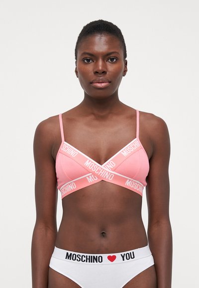 Rózsaszín bralette vékony pántokkal és keresztbe fonott dizájnnal, fehér "MOSCHINO" logós pánttal. Fehér alsóneművel kombinálva.
