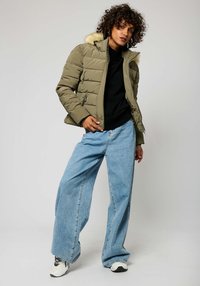 Olivengroene pufferjack met een nep bont capuchon, ritszakken en een gestikt ontwerp, gecombineerd met lichtblauwe wijde jeans en sneakers.