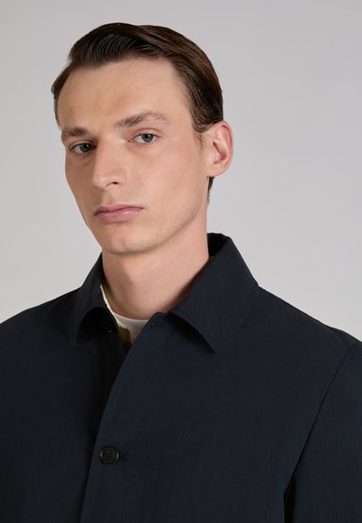 Jeune homme aux cheveux bruns coiffés en arrière porte un manteau à boutons foncé sur une chemise claire, regardant légèrement vers le bas avec une expression neutre.