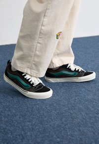 Černé a hnědé tenisky s tyrkysovými pruhy, bílé tkaničky a bílá gumová podrážka, v kombinaci s krémově barevnými pohodlnými kalhotami.