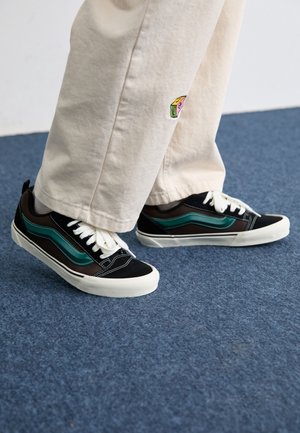 Baskets noires et marron avec des rayures turquoise, lacets blancs et semelle en caoutchouc blanche, associées à un pantalon crème décontracté.