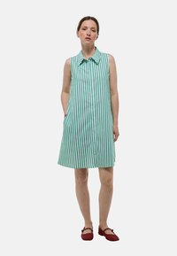 Robe sans manches avec des rayures verticales vertes et blanches, devant à boutons, col et poches latérales ; assortie avec des chaussures plates rouges.