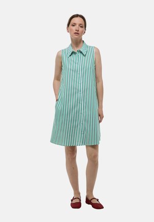Robe sans manches avec des rayures verticales vertes et blanches, devant à boutons, col et poches latérales ; assortie avec des chaussures plates rouges.