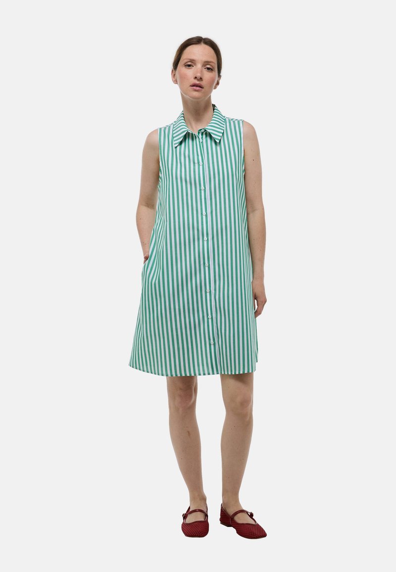 Robe sans manches avec des rayures verticales vertes et blanches, devant à boutons, col et poches latérales ; assortie avec des chaussures plates rouges.