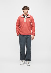 Νέος άνδρας που φοράει κρεμώδες κοραλλί πουκάμισο από φλις, σκούρο γκρι παντελόνια και λευκά αθλητικά παπούτσια, στέκεται μπροστά σε απλό φωτεινό φόντο, κοιτάζοντας πλάγια.