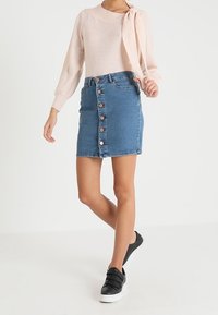 Heller rosa Pullover mit langen Ärmeln und Schleifendetail, kombiniert mit einem blauen Jeansrock mit Knöpfen vorne und schwarzen Slippers.