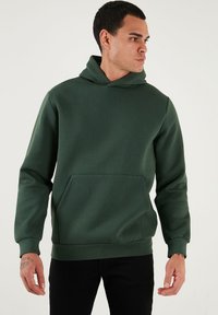 Tmavozelená mikina vyrobená z jemnej, hladkej látky. Obsahuje klokanie vrecko a rebrované manžety, so sťahovacou kapucňou a voľným strihom.