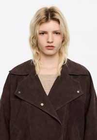 Bruine suède oversized jas met een omslagkraag, zilveren drukknoopjes en zijzakken. Draag het over een lichtbeige gebreide trui.