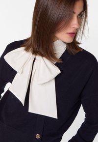 Marineblauer Cardigan mit einer großen weißen Schleife am Kragen. Hat eine weiche Textur und einen goldenen Logo-Button auf der Vorderseite.