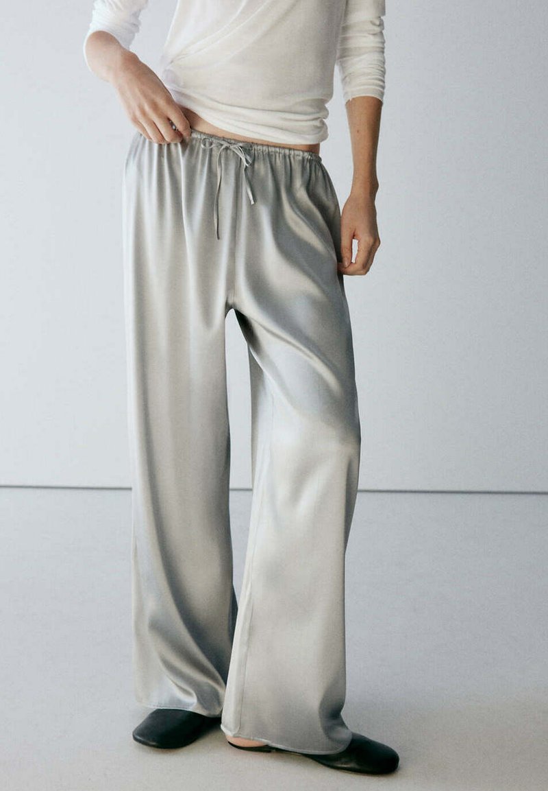 Personne portant un pantalon ample en satin gris brillant avec une taille à cordon et des chaussures noires sans lacets, debout devant un fond clair uni.