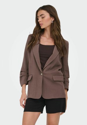 Nő barna blézerben sötét felsővel és fekete rövidnadrággal, enyhén oldalra fordított fejjel, fehér háttér előtt áll.