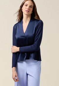 Mujer de cabello largo que lleva pendientes geométricos dorados, blusa de raso azul marino y pantalones azul claro, de pie frente a un fondo beige.