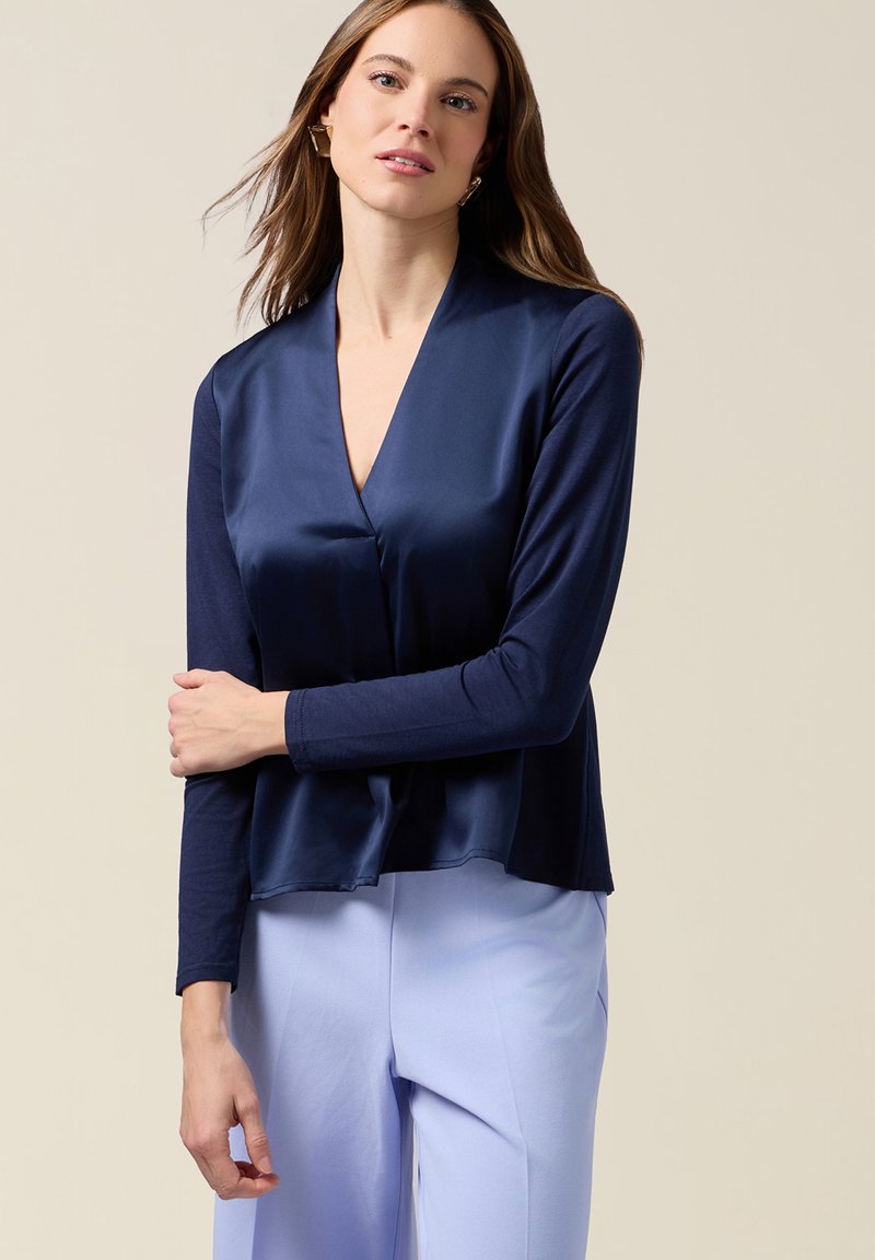 Mujer de cabello largo que lleva pendientes geométricos dorados, blusa de raso azul marino y pantalones azul claro, de pie frente a un fondo beige.