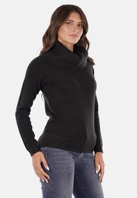 Maglione nero lavorato a maglia con collo a barchetta e polsini a costine. La texture è liscia con una leggera lucentezza, abbinato a jeans aderenti.