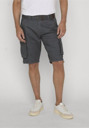 Mann trägt dunkelgraue Cargo-Shorts mit einem schwarzen Gürtel, weiße Sneaker und ein weißes Hemd, steht vor einem einfarbigen Hintergrund.
