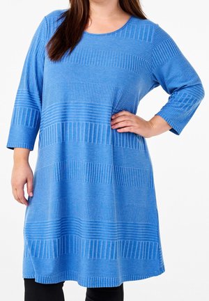 Žena v modrých šatech po kolena s dlouhým rukávem a střídavými vodorovnými texturovanými pruhy pózuje s jednou rukou v bok.