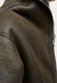 Bruine nepleer jas met een gladde textuur, hoge kraag en zilveren ritssluiting. Toont de schoudernaad en de binnenvoering.