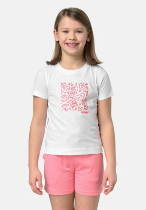 Ragazza sorridente con una maglietta bianca con motivo floreale rosa e pantaloncini rosa, in piedi davanti a uno sfondo chiaro e semplice.