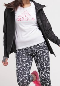 Musta urheiluvesti, valkoinen T-paita, jossa on vaaleanpunainen Adidas-logo, kuviolliset mustat legginsit ja vaaleanpunaiset juoksukengät. Harmaa teksturoitu rannekoru.