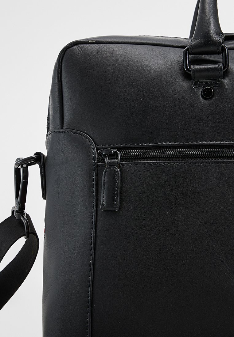 Malette en cuir noir avec poche avant zippée et sangle d'épaule ajustable, sur un fond uni.
