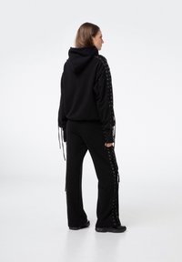 Czarna bluza z kapturem z bocznymi sznurkami i dopasowanymi spodniami. Miękka tkanina, luźny krój oraz zwisające troczki na rękawach i nogawkach.