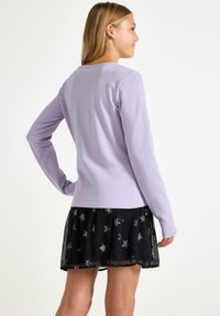 Kvinde med blond hår iført en lys lilla langærmet skjorte og en sort nederdel med sølvstjerne-mønster, der står foran en hvid baggrund.