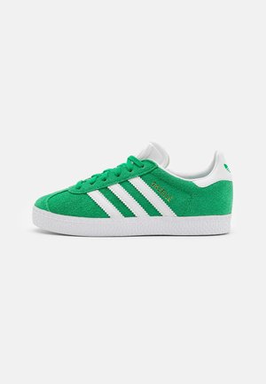Grøn ruskinds-sneaker med hvid sål, hvide Adidas-striber og "Gazelle" tekst i guld på siden.