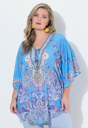 Blonditukkainen nainen, jolla on keltaiset kukka-aiheiset korvakorut ja sininen, kukkakuvioinen väljä paita, jossa on 3/4-hihat.