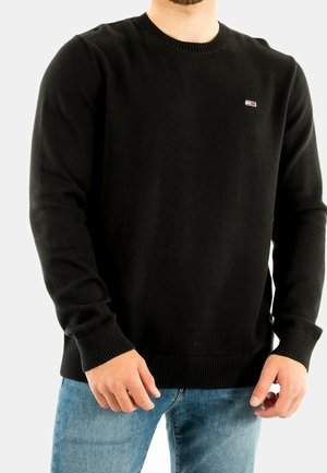 Mann trägt schwarzen langärmligen Pullover mit Rundhalsausschnitt und kleinem Logo auf der Brust sowie blaue Jeans, steht vor einem einfarbigen Hintergrund.