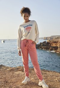 Lichtbeige sweatshirt met "Sunrise" graphic en roze sweatpants met een elastische trekkoord tailleband, gecombineerd met witte sportschoenen.