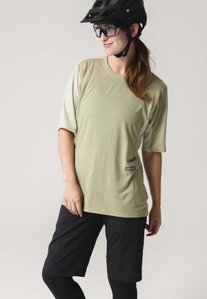 Frau mit schwarzem Fahrradhelm, Schutzbrille, hellgrünem kurzärmligem Shirt, schwarzer Shorts und schwarzen Leggings, die vor einem schlichten Hintergrund steht.