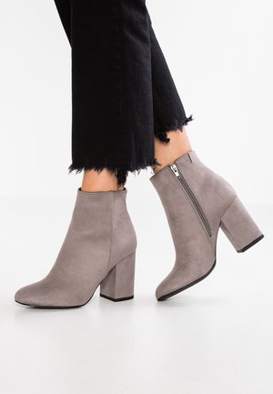 Bottes bottines en daim gris avec talons carrés et fermetures éclair latérales portées avec un pantalon noir à ourlet effiloché sur un sol blanc.