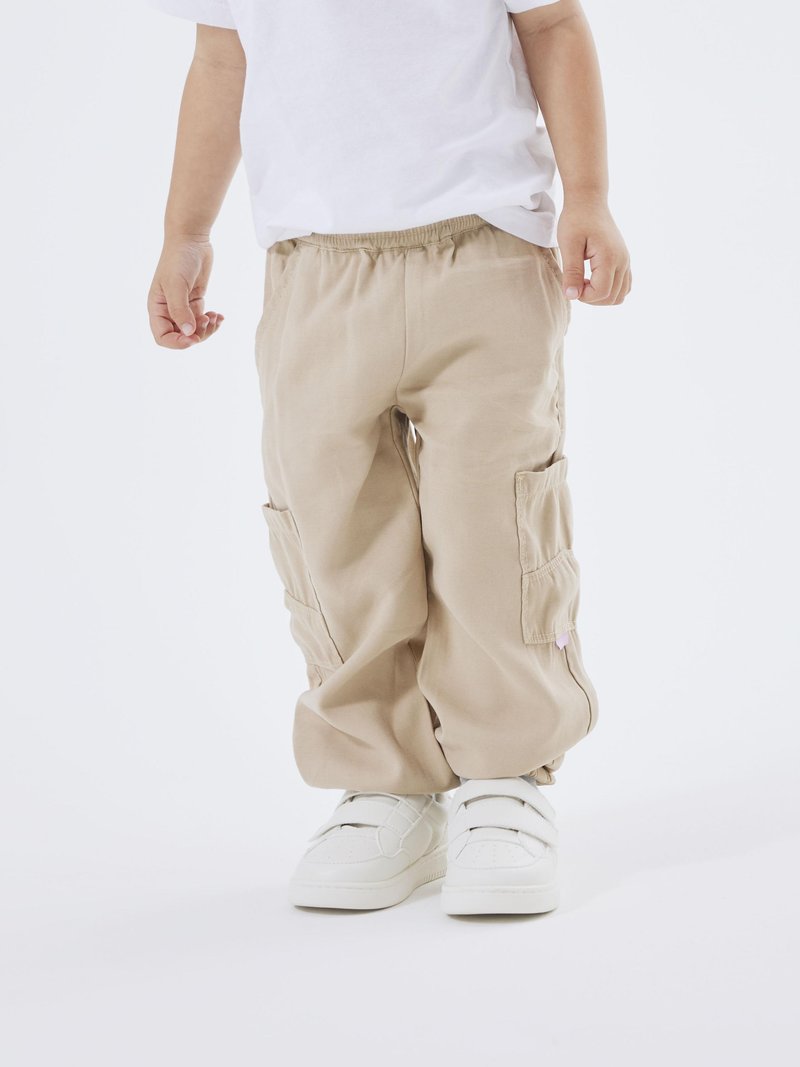 Pantalones cargo beige con cinturilla elástica y bolsillos laterales, combinados con zapatillas blancas con correas de velcro.