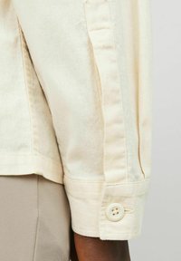Chemise beige clair à manches longues avec des poignets à bouton, présentant une texture subtile et un design classique. Couture visible le long de la couture.