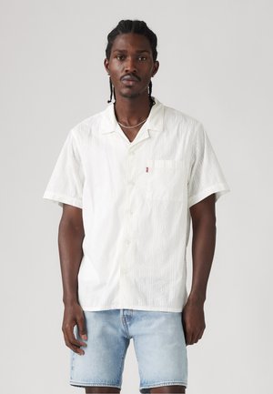Short-sleeve white shirt with a textured pattern, featuring a chest pocket and large buttons, paired with light blue denim shorts.