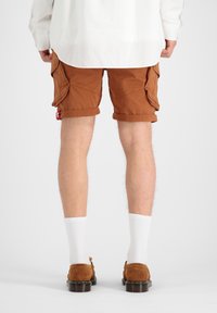 Shorts cargo marron avec des ourlets retroussés, dotés de deux poches latérales et d'une petite étiquette rouge. Portés avec des chaussettes blanches et des chaussures en daim marron.