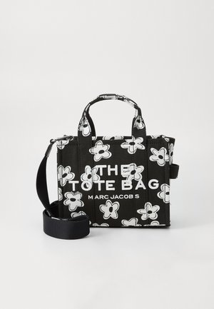 Bolso tote negro con patrón floral blanco, que cuenta con dos asas superiores y una correa ajustable y desmontable. El texto dice "EL BOLSO TOTE" y "MARC JACOBS."