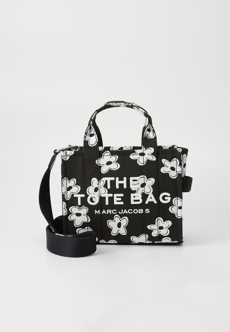 Sac fourre-tout noir avec un motif floral blanc, doté de deux poignées supérieures et d'une sangle ajustable et amovible. Le texte indique "LE SAC FOURRE-TOUT" et "MARC JACOBS."