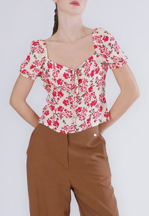 Mujer con blusa crema con estampado floral rojo y mangas abullonadas, combinada con pantalones marrones de cintura alta, con las manos apoyadas en las caderas.