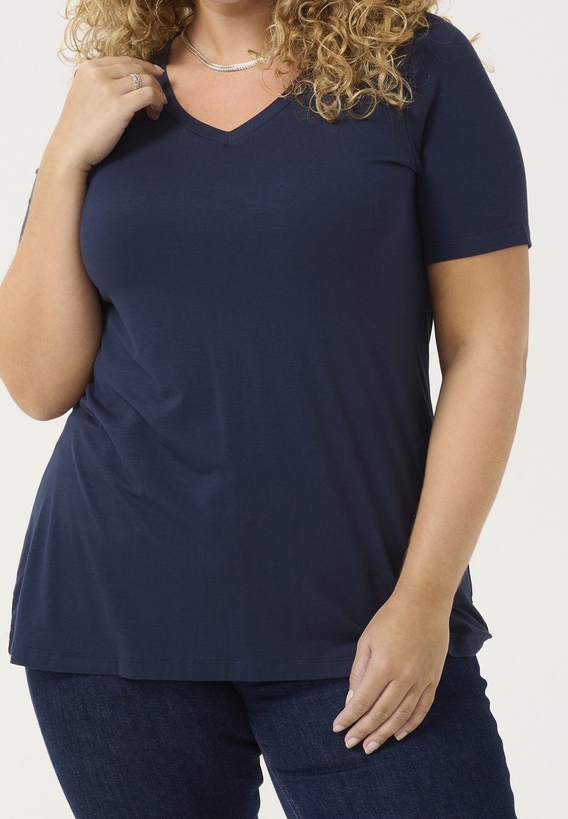 Personne aux cheveux bouclés portant un t-shirt bleu marine à manches courtes en V et un jean foncé, avec un collier et une bague en argent, la main posée près de la clavicule.
