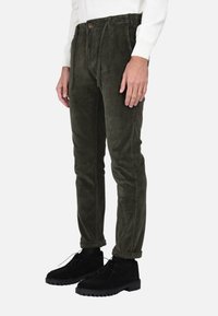 Pantaloni in corduroy verde oliva con una vestibilità comoda, dotati di un laccetto, tasche frontali e chiusura a bottone. Indossati con stivali neri.