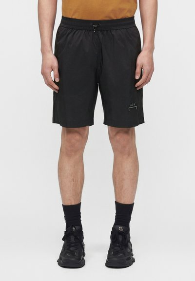 Homme portant un short de sport noir avec une taille à cordon, des chaussettes noires montantes et des baskets noires, debout devant un fond uni.