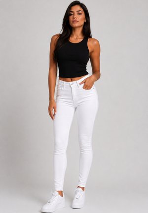 Kvinde iført sort ærmeløs crop top, hvide højtaljede skinny jeans og hvide sneakers, stående mod en grå baggrund.