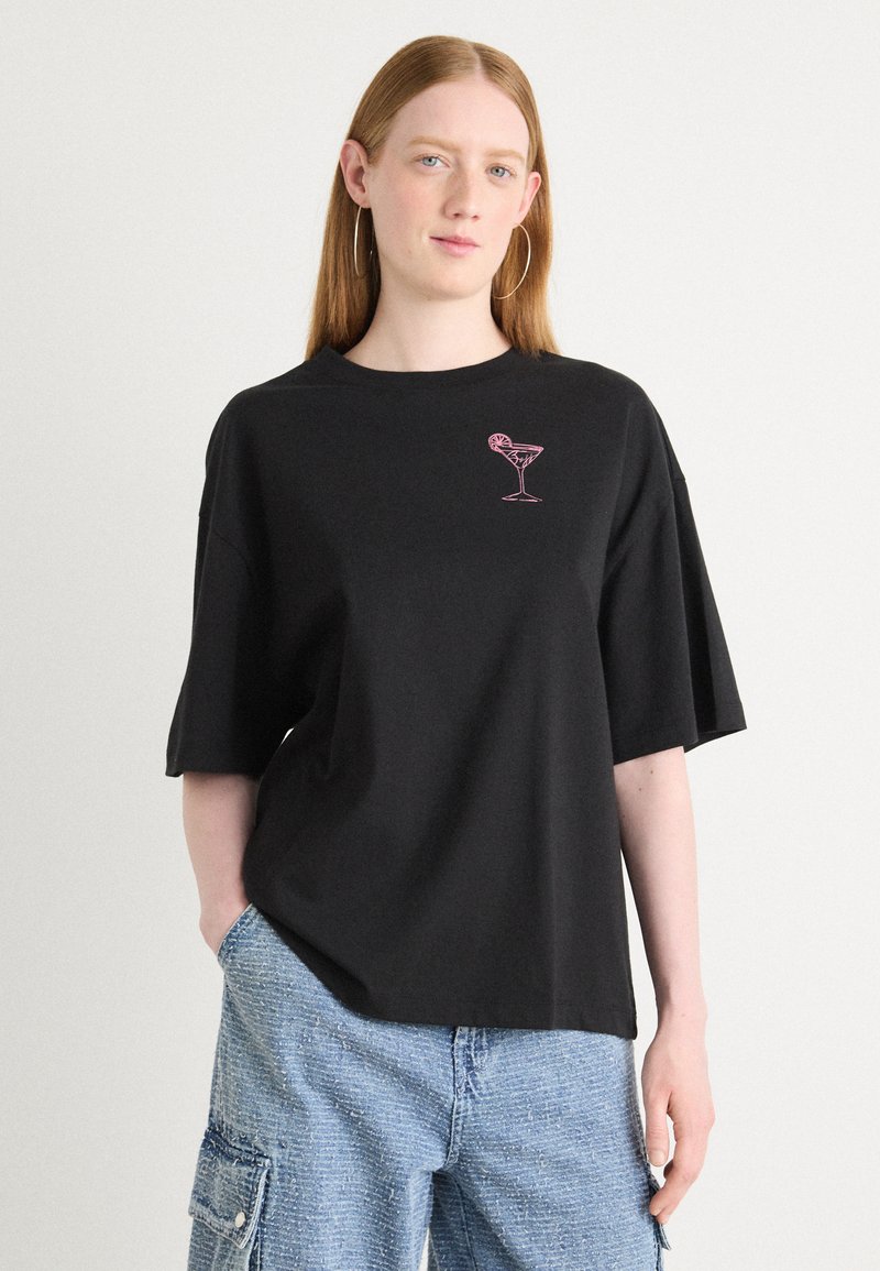 Black oversized t-shirt featuring a pink cocktail glass graphic on the left chest. The material appears smooth, with short sleeves.