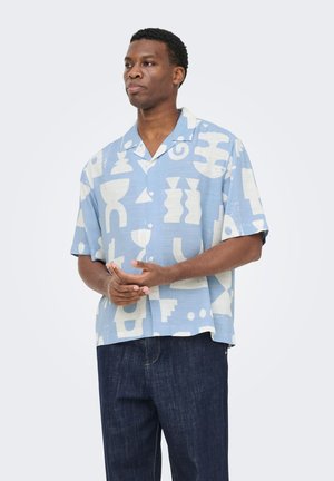 Muž v svetlomodrej košeli s krátkym rukávom s bielymi abstraktnými tvarmi a tmavomodrých džínsoch, stojaci so zloženými rukami.