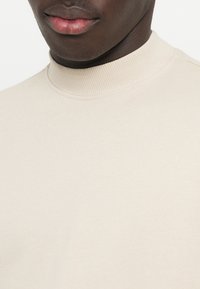 Maglione beige con collo alto realizzato in tessuto morbido, caratterizzato da un colletto a coste e una superficie liscia, con un design che punta sulla semplicità.