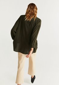 Blusa verde oliva de corte holgado, con manga larga y puños abotonados, combinada con pantalones de pierna ancha color crema y zapatos negros.