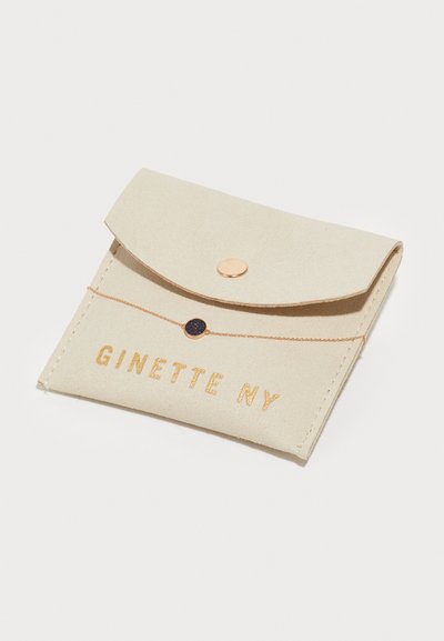 Béžový filcový držiteľ na karty s preklopným uzáverom, zlatým patentným gombíkom a embosovaným textom "GINETTE NY". Jednoduchý tvar s prešívaním.