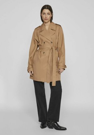 Gabardina beige con frente de doble botonadura, cinturón en la cintura y acentos de botones; llevada sobre pantalones negros de pierna ancha y botas negras de punta.